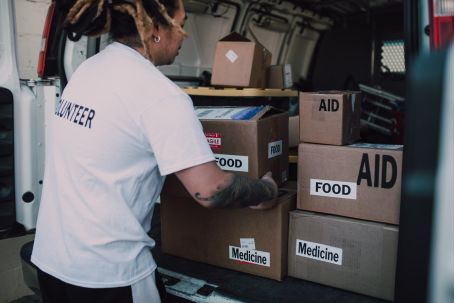 Man in White T-shirt and Gray Pants Standing Beside Brown Cardboard Boxes